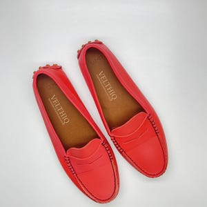 Può includere: Un paio di mocassini in pelle rosso vivo. Le scarpe hanno un design classico con tomaia cucita e soletta marrone. Il marchio "VELTHIQ" è stampato sulla soletta. Taglia 37.