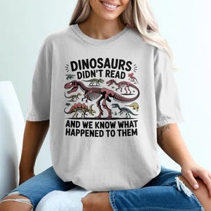May include: White t-shirt with a dinosaur-themed graphic. The design features various dinosaur illustrations and the text "DINOSAURS DIDN'T READ AND WE KNOW WHAT HAPPENED TO THEM" in black. The shirt is worn by a person.