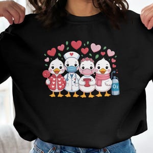 May include: Black sweatshirt featuring a cartoon graphic of four ducks dressed as healthcare workers, each with unique accessories like a lollipop, stethoscope, and oxygen tank. Pink and red hearts are above the ducks.