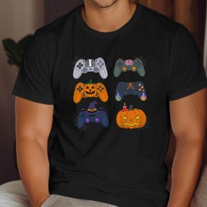 Op de afbeelding: Zwart T-shirt met zes Halloween-thema's voor gamecontrollers. De controllers hebben ontwerpen zoals een skelet, hersenen, pompoen en heksenhoed. De oranje en paarse controllers hebben een pompoenthema. Het shirt is gemaakt van zacht materiaal.