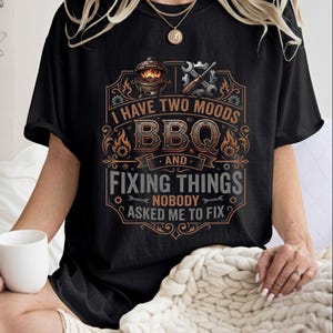 May include: A black short-sleeved t-shirt featuring a graphic print with the text I HAVE TWO MOODS BBQ AND FIXING THINGS NOBODY ASKED ME TO FIX. The design includes illustrations of a charcoal grill, a wrench, and a screwdriver with gear accents.