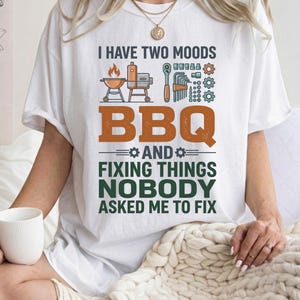 May include: A white t-shirt featuring a graphic print with the text I HAVE TWO MOODS BBQ AND FIXING THINGS NOBODY ASKED ME TO FIX. The design includes illustrations of a grill, a smoker, a ratchet, hex keys, and various metal gears in orange, green, and grey.