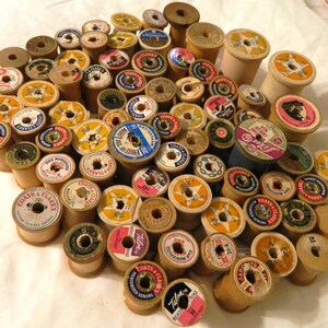 Pode incluir: Uma coleção de bobinas de madeira vintage, muitas com etiquetas coloridas. As bobinas variam em tamanho e estão dispostas sobre uma superfície branca. Os rótulos incluem texto como "Coats & Clark" e "Talon".