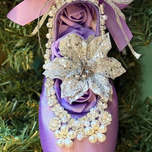May include: A decorative purple satin ballet slipper ornament. The slipper is adorned with a silver glitter flower, faux pearls, and a matching purple bow. The interior is filled with purple fabric roses, creating a festive holiday decoration.