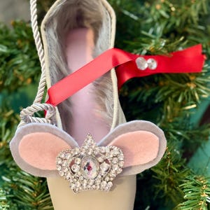 May include: A decorative ballet slipper ornament with a mouse-ear design and a jeweled crown. The slipper is beige with a pink interior and a red ribbon. The ornament is hanging from a silver rope and is set against a backdrop of green pine needles.