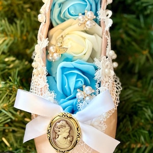 May include: A decorative ballet slipper filled with blue and white roses. The slipper is adorned with white lace, pearls, and a gold cameo. A white ribbon and bow are attached to the front, creating an elegant and romantic aesthetic.