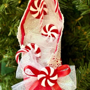 May include: A festive Christmas ornament shaped like a shoe, adorned with red and white peppermint candies and candy canes. The shoe is red with white lace trim and a white tulle bow. The ornament is set against a backdrop of green Christmas tree needles.