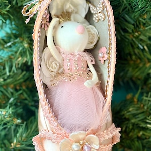 May include: A decorative ornament shaped like a ballet slipper, featuring a white mouse in a pink tutu. The slipper is adorned with lace, pearls, and a floral embellishment. The background includes greenery.