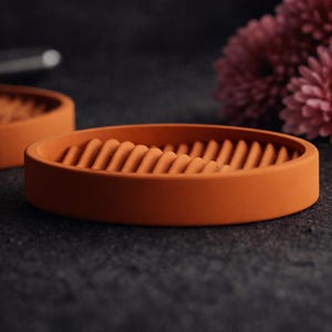 Könnte beinhalten: Eine orangefarbene Seifenschale mit geriffeltem Innenraum. Die runde Schale besteht aus einem matten Material und ist für die Aufnahme eines Seifenstücks konzipiert. Der Hintergrund ist dunkel, mit einem verschwommenen Objekt und Blumen.