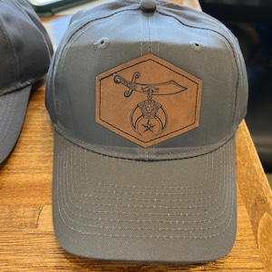 May include: Gray baseball cap with a brown leather patch featuring a sword, crescent moon, and star design. The cap has a curved brim and a button on top. The hat is made of a durable fabric.