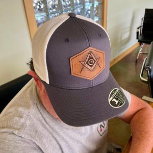 May include: A charcoal gray and white trucker hat with a leather patch featuring a Masonic symbol. The patch is brown with a compass, square, and the letter "G". The hat has a curved brim and a Flexfit 110 sticker.