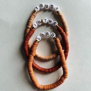 May include: Three stacked bracelets with a neutral color palette. The bracelets are made of small, flat, round beads in shades of tan, brown, and orange. White letter beads spell out "TRUST IN THE LORD" on each bracelet. Gold accents are visible.