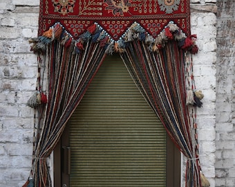 Op de afbeelding: Een decoratieve deurafdekking met een rode tapijt header met bloemen en geometrische patronen. Het tapijt is versierd met kleurrijke kwasten en leidt naar gestreepte gordijnen. De gordijnen zijn vastgebonden met kwasten, waardoor een uniek deuropeningontwerp ontstaat.