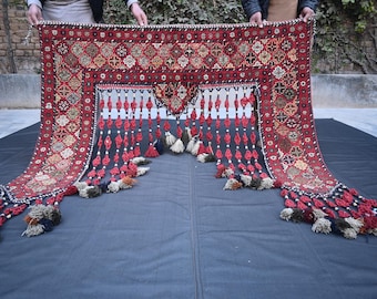 Op de afbeelding: Een decoratief textiel in rood en zwart, met ingewikkelde geometrische patronen en hangende kwasten. Het textiel heeft een centraal ontwerp met een rand van herhalende vormen en wordt door twee personen vastgehouden.