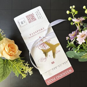 May include: A wedding invitation designed as a passport, featuring a gold airplane charm and a white ribbon. The invitation includes the text "PASSPORT WEDDING INVITATION" and the date "06.09.2025".