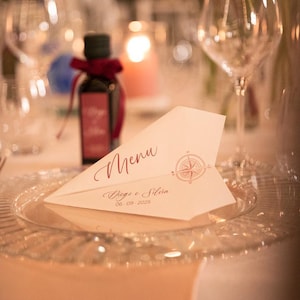 Puede incluir: Un menú en forma de avión de papel con la palabra "Menu" en escritura cursiva y un diseño de rosa de los vientos. La tarjeta está sobre un plato de cristal transparente, con una botella pequeña y copas de vino al fondo. La fecha "06-09-2025" está impresa en la tarjeta.