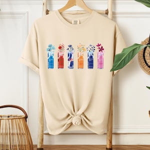 K&ouml;nnte beinhalten: Beigefarbenes T-Shirt mit einem Aquarelldesign von sechs Getr&auml;nkedosen, die jeweils eine andere Anordnung bunter Blumen enthalten. Die Dosen sind in Blau-, Rot-, Orange- und Rosat&ouml;nen gehalten, mit floralen Akzenten.