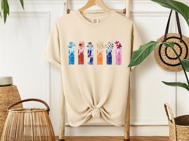 K&ouml;nnte beinhalten: Beigefarbenes T-Shirt mit einem Aquarelldesign von sechs Getr&auml;nkedosen, die jeweils eine andere Anordnung bunter Blumen enthalten. Die Dosen sind in Blau-, Rot-, Orange- und Rosat&ouml;nen gehalten, mit floralen Akzenten.