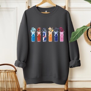 K&ouml;nnte beinhalten: Dunkelgraues Sweatshirt mit einem Aquarelldesign von sechs bunten Getr&auml;nkedosen, die jeweils eine andere Blumenanordnung enthalten. Die Dosen sind in Blau-, Rot-, Wei&szlig;-, Orange- und Rosat&ouml;nen gehalten und mit Schleifen versehen. Ein l&auml;ssiges Kleidungsst&uuml;ck mit floralem Thema.