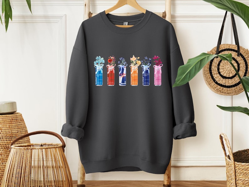 K&ouml;nnte beinhalten: Dunkelgraues Sweatshirt mit einem Aquarelldesign von sechs bunten Getr&auml;nkedosen, die jeweils eine andere Blumenanordnung enthalten. Die Dosen sind in Blau-, Rot-, Wei&szlig;-, Orange- und Rosat&ouml;nen gehalten und mit Schleifen versehen. Ein l&auml;ssiges Kleidungsst&uuml;ck mit floralem Thema.