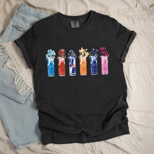 K&ouml;nnte beinhalten: Schwarzes T-Shirt mit einem farbenfrohen Blumendesign. Die Grafik zeigt sechs stilisierte Vasen, jede gef&uuml;llt mit verschiedenen Blumen und mit einem Band gebunden. Auf der linken Seite des Bildes ist eine hellblaue Jeans zu sehen.