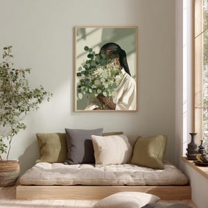 May include: A framed art print featuring a person holding a bouquet of green and white flowers. The person is wearing a white shirt and gold hoop earrings. The artwork has a neutral color palette and is displayed on a light green wall.