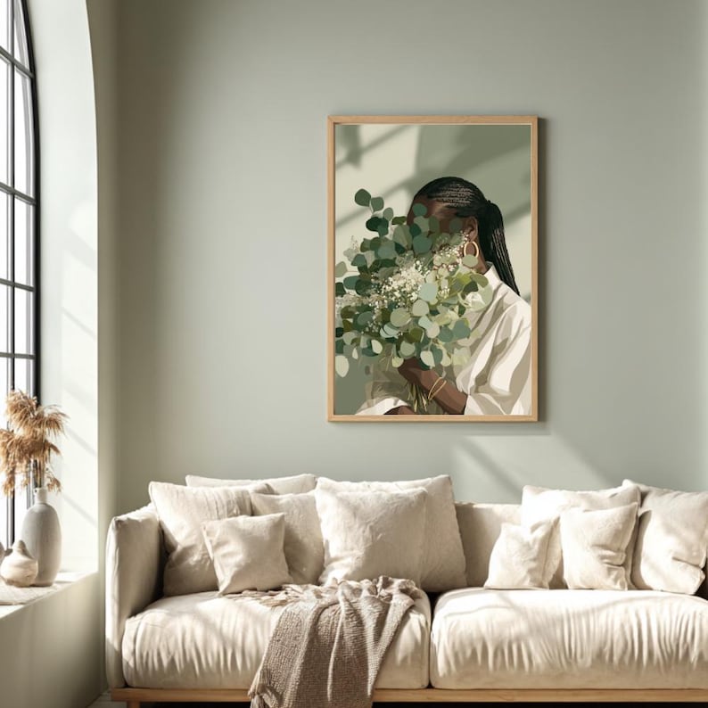 May include: A framed art print featuring a person holding a bouquet of green and white flowers. The person is wearing a white shirt and gold hoop earrings. The artwork is displayed on a light green wall above a beige sofa.