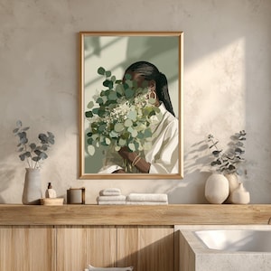 May include: A framed art print featuring a person holding a bouquet of green and white flowers. The person is wearing a white shirt and gold hoop earrings. The artwork has a neutral color palette and is displayed in a bathroom setting.