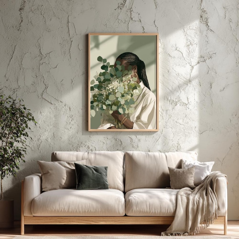 May include: A framed art print depicts a person holding a bouquet of green and white flowers, partially obscuring their face. The person wears a white shirt and gold hoop earrings. The artwork is displayed above a beige sofa with pillows and a blanket.