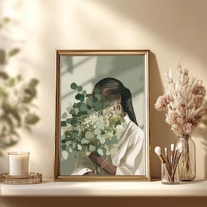 May include: A framed art print featuring a person holding a bouquet of green and white flowers, with a gold frame. The person is wearing a white shirt and gold hoop earrings. A candle and decorative vase are also visible.