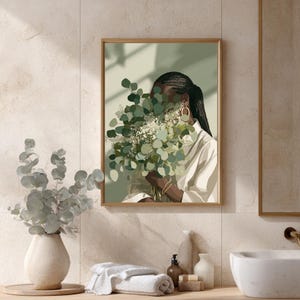 May include: A framed art print featuring a person holding a bouquet of green and white flowers. The person is wearing a white shirt and gold hoop earrings. The artwork has a soft, muted color palette with a gold frame.