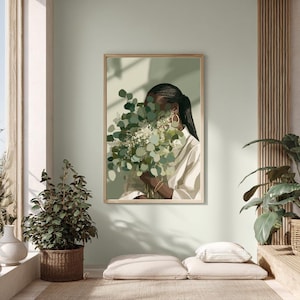 May include: A framed art print featuring a person holding a bouquet of green and white flowers, with a light green background. The person is wearing a white shirt and gold hoop earrings. The artwork is displayed in a well-lit room with plants.