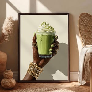 Puede incluir: Impresión artística enmarcada que muestra una mano sosteniendo un matcha latte. La mano está adornada con anillos y pulseras doradas, y las uñas están pintadas de verde. La bebida está cubierta con crema batida, sobre un fondo beige neutro.