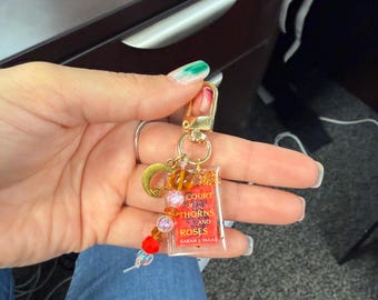 May include: A gold-colored keychain with a charm featuring a miniature book with the title "Court of Thorns and Roses" and the author's name. The keychain also includes a crescent moon charm and colorful beads.