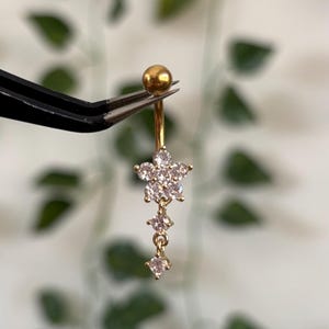 Pode incluir: Um piercing de umbigo dourado com um design floral. A bola superior é segurada por uma pinça. O design floral apresenta um aglomerado de pequenas pedras preciosas transparentes em forma de estrela, com três pedras preciosas penduradas abaixo.