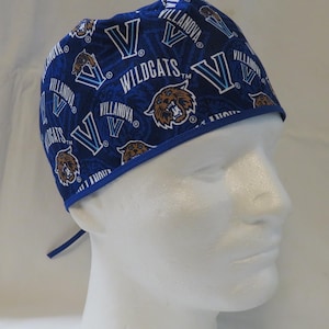 May include: Blue Villanova Wildcats scrub cap with a repeating pattern of the team logo, the letter "V", and the word "Villanova". The cap has a blue trim and ties in the back. The cap is on a white mannequin head.