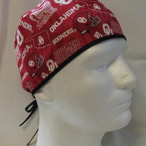 May include: A red and white patterned cap featuring the words "Oklahoma Sooners" and various related graphics. The cap has a black trim and black ties for securing. The design includes football helmets and the state outline.