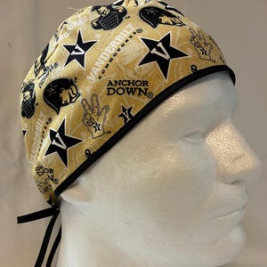 May include: A patterned scrub cap featuring the text "Vanderbilt Commodores" and "Anchor Down" in black on a gold background. The cap has black trim and ties.