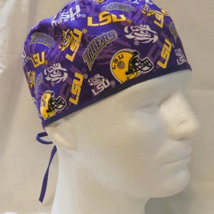 May include: Purple and gold LSU Tigers scrub cap. The cap features a repeating pattern of the LSU logo, tiger eyes, and football helmets. The cap has a dark purple trim and ties.