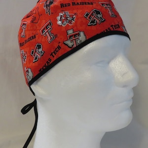May include: An orange scrub cap featuring the words "Red Raiders" and "Texas Tech" with various logos. The cap has a black trim and ties in the back.