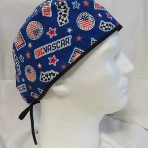 May include: Blue surgical scrub hat with a black trim. The hat has a pattern of red, white, and blue stars, checkered flags, and the words "United States of America" and "NASCAR".