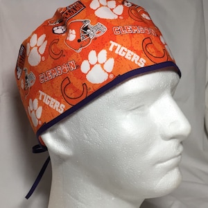 May include: Orange surgical scrub cap with a repeating pattern of white paw prints, football helmets, and the words "CLEMSON" and "TIGERS." The cap has a dark purple trim and ties.