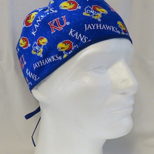 May include: A blue surgical scrub cap featuring the University of Kansas Jayhawks logo. The cap has a repeating pattern of the Jayhawk mascot, the letters "KU", and the word "JAYHAWKS" in white. The cap has a dark blue trim and ties.