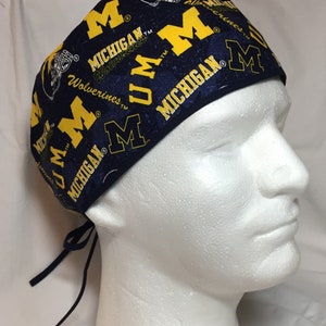 May include: A navy blue and yellow patterned scrub cap featuring the text "MICHIGAN Wolverines" and the letter "M". The cap has a tie closure at the back and is displayed on a white mannequin head.