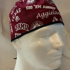 May include: Maroon and white patterned scrub cap featuring the text "Gig 'Em Aggies" and "Aggieland". The cap has a black trim and is designed to be worn on the head. The cap is made of a soft, comfortable material.