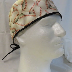 May include: A white scrub hat with a black trim and a baseball pattern. The hat has a black tie that can be tied at the back of the neck.