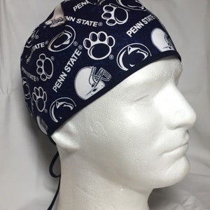 May include: Navy blue surgical scrub cap with white Penn State logos, paw prints, and football helmets. The cap has a tie in the back for an adjustable fit. The fabric has a subtle, mottled texture.