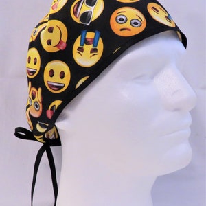 May include: Black scrub hat with a yellow and black emoji pattern. The hat has black ties.
