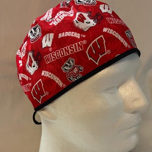 May include: A red and white patterned scrub cap featuring the Wisconsin Badgers logo and text. The cap has a black trim and a tie in the back. The design includes the word "Badgers" and the letter "W".