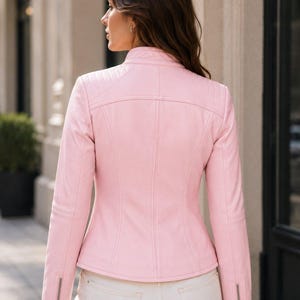 Puede incluir: Una chaqueta de color rosa claro con cuello alzado, paneles acolchados en los hombros y mangas largas con cremalleras met&aacute;licas plateadas en los pu&ntilde;os. La prenda presenta costuras verticales en la espalda y se combina con pantalones claros.